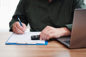 Person filling out paperwork with a car key on the table next to a laptop, preparing to sell a Nissan in California.